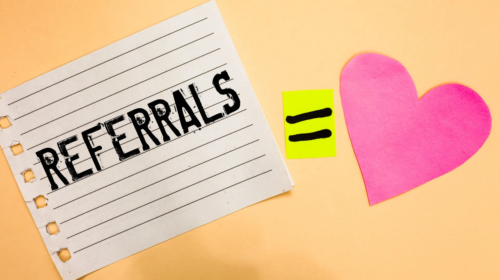 A piece of paper with the word 'REFERRALS' written on it, an equal sign, and a pink heart symbol on a light orange background