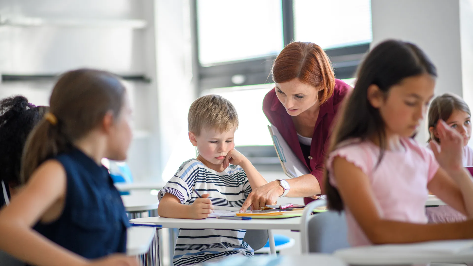 A teacher assisting a young student in a classroom with other children