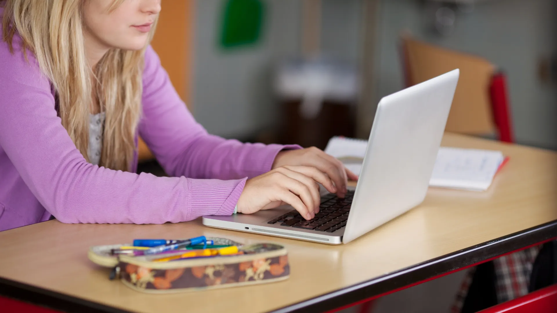 A student using a laptop at a desk with a pencil case and pens nearby.