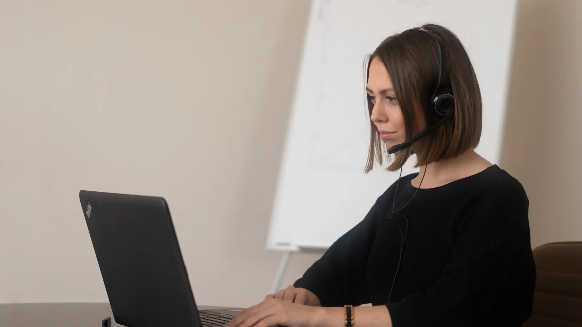A person working on a laptop while wearing a headset, with a whiteboard in the background