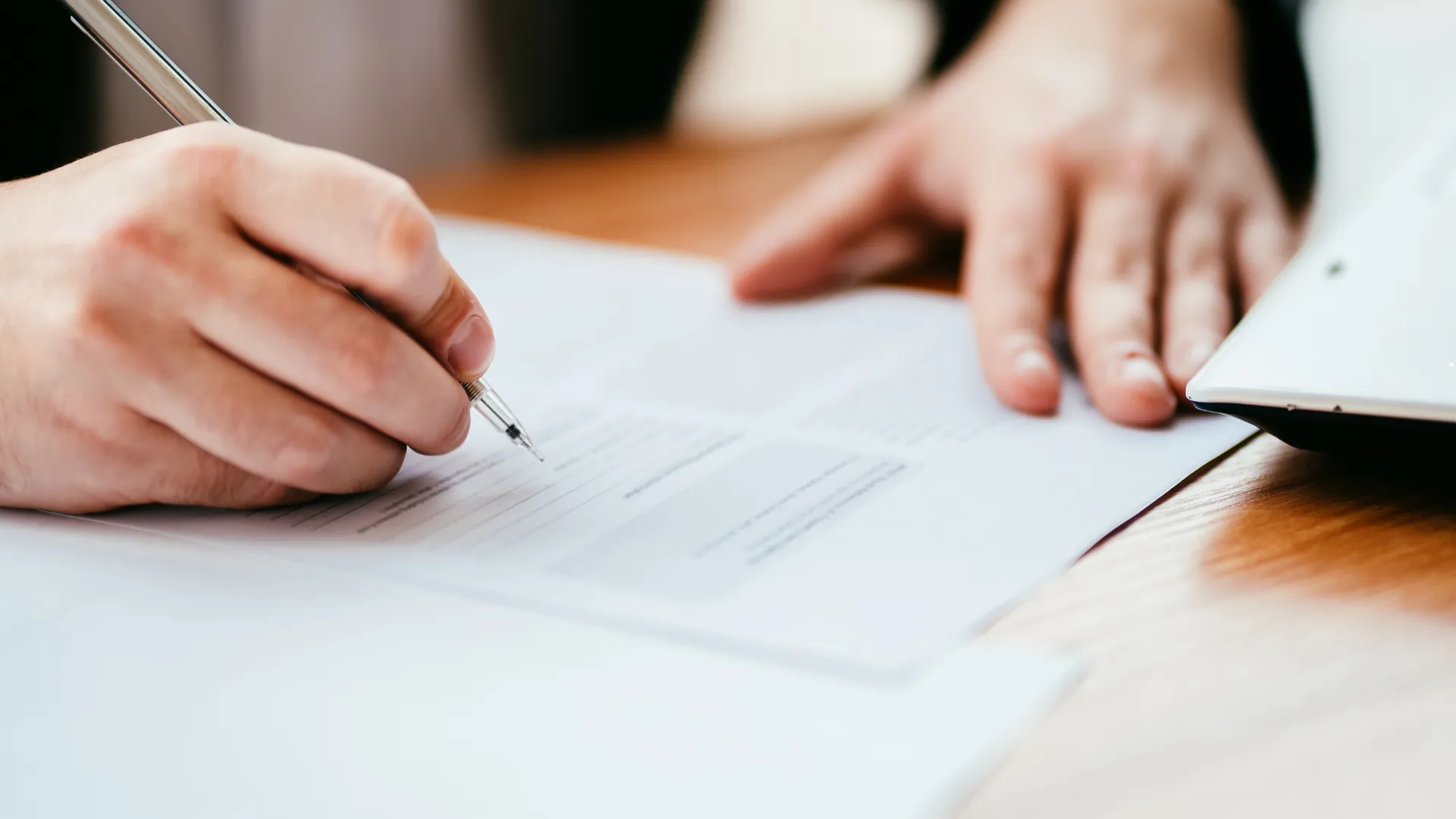 A person writing on a document with a pen, with a laptop partially visible on the side