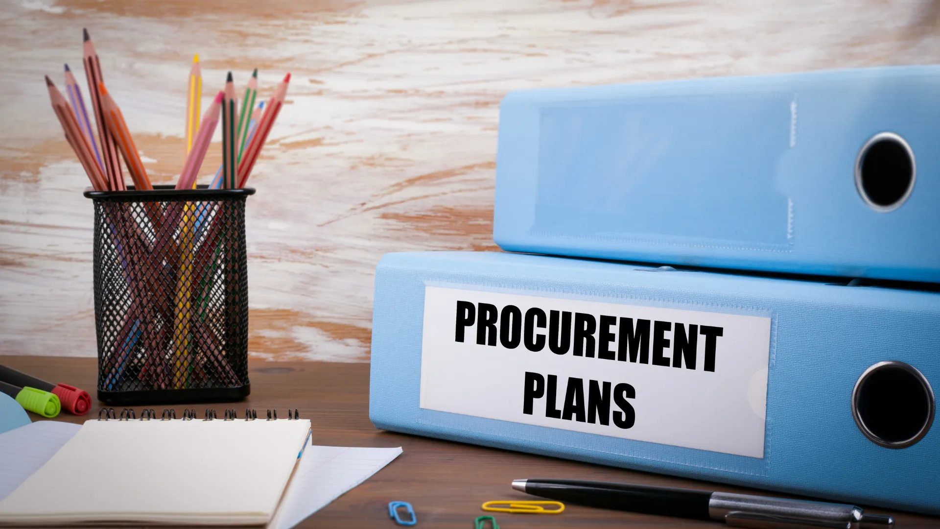 Two blue binders labelled "PROCUREMENT PLANS" on a desk with office supplies.