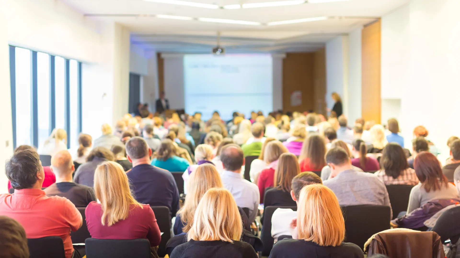 A large group of people seated in a conference room, facing a presentation screen at the front