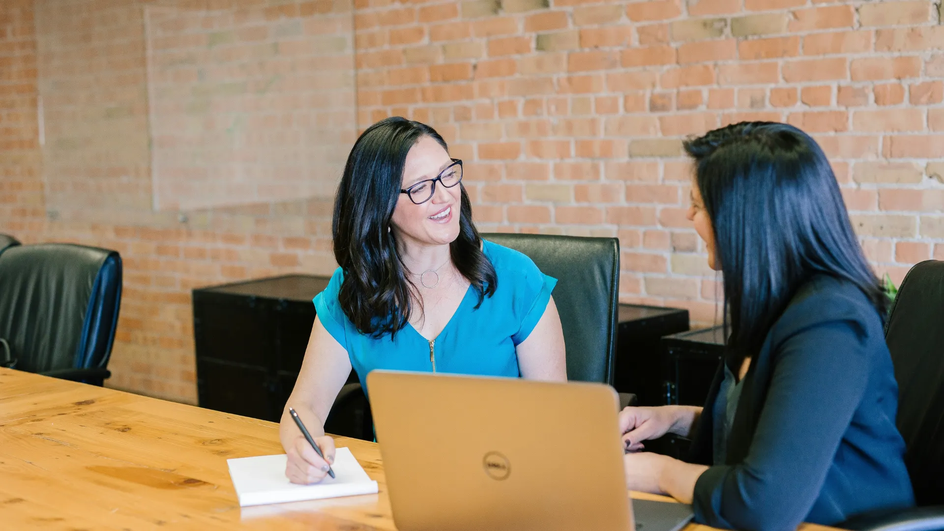 Two people in a meeting seated at a wooden table. One person is writing on a notepad while the other is using a laptop