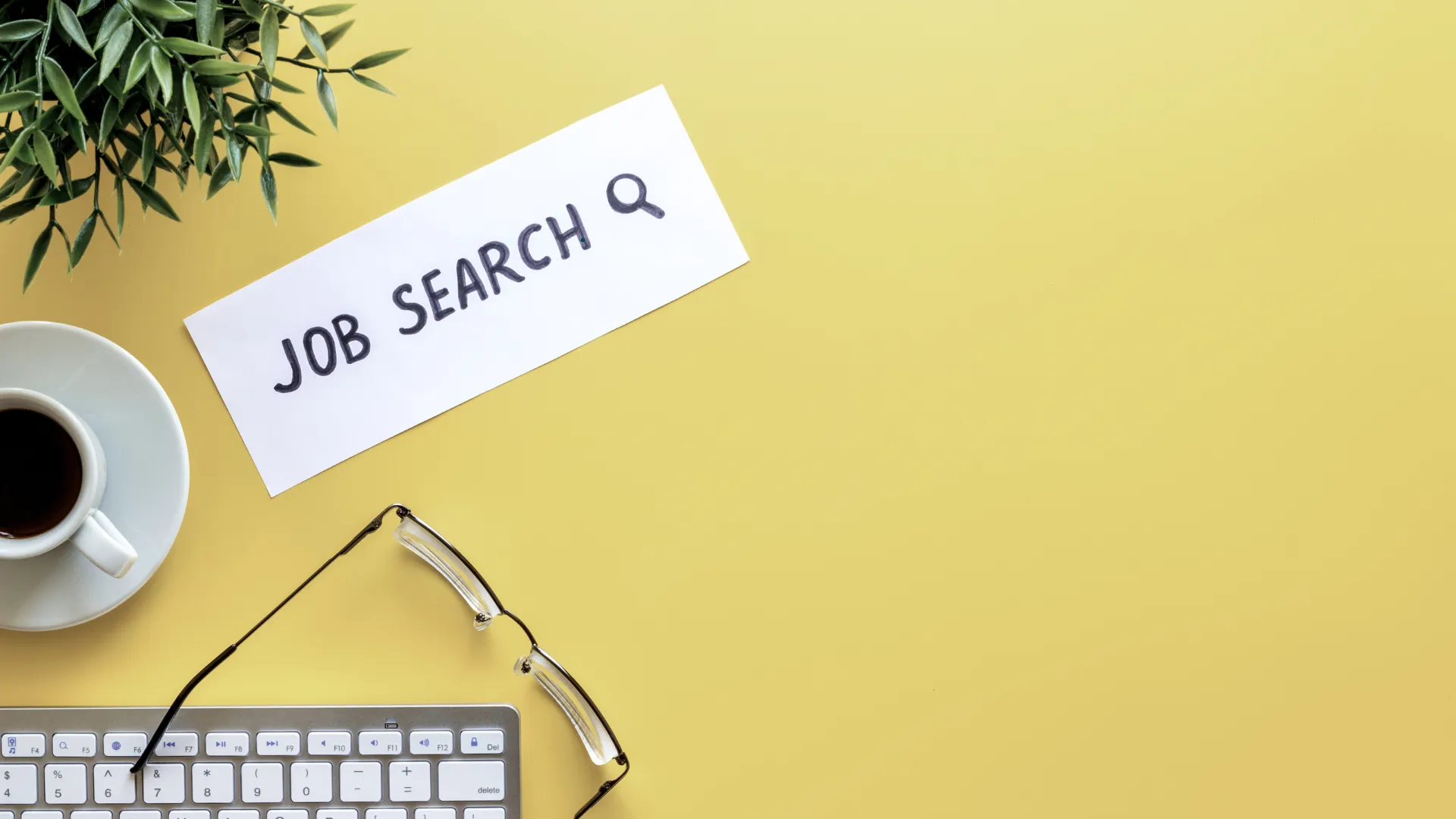 A workspace with a keyboard, coffee, glasses, and a paper labelled "JOB SEARCH".