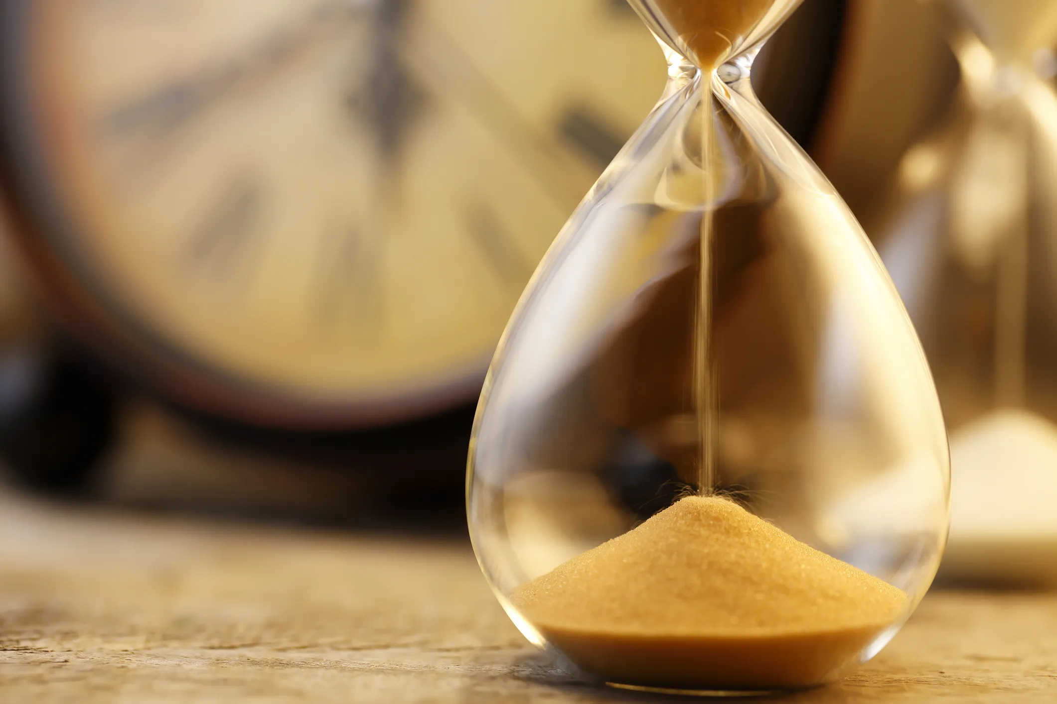 A close-up of an hourglass with sand flowing into the bottom chamber, with a blurred clock face in the background.