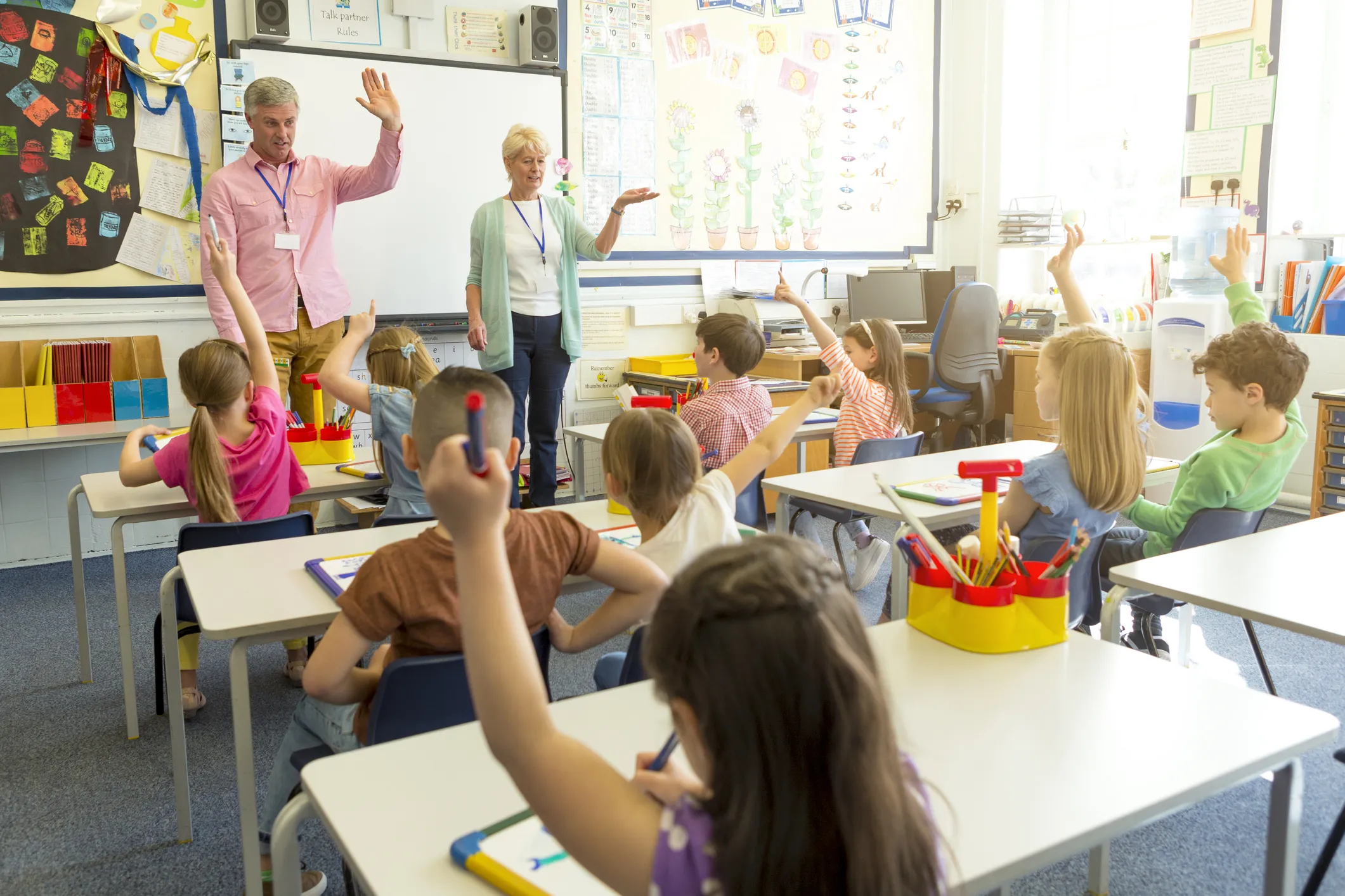 A classroom with children seated at desks raising their hands, while two adults stand at the front leading the lesson