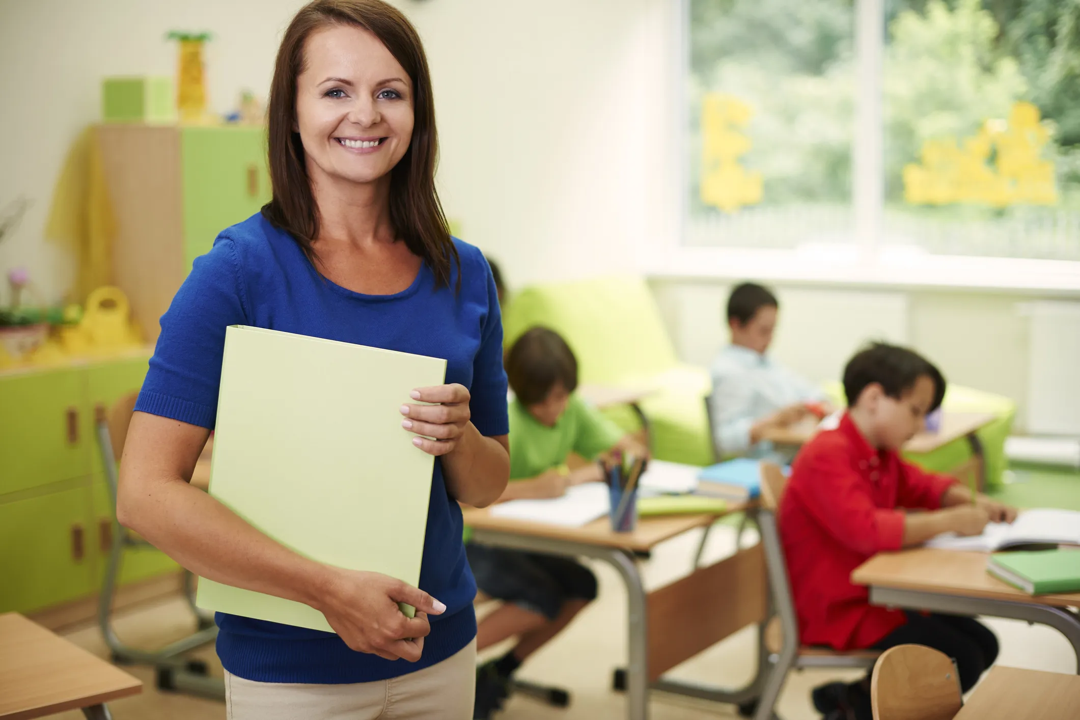 Teacher holding a folder in a classroom with children working at desks.