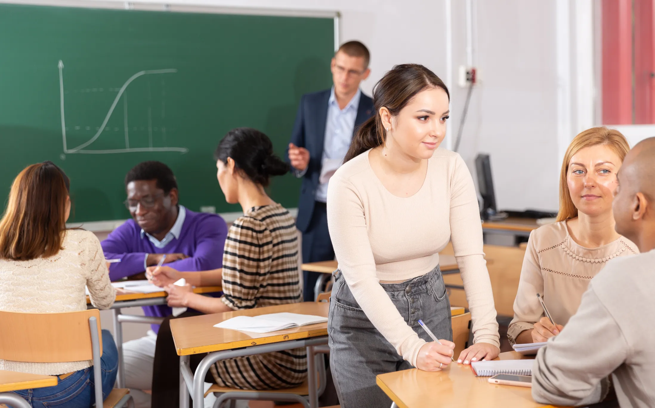 Students in a classroom working in small groups while a teacher stands near a chalkboard