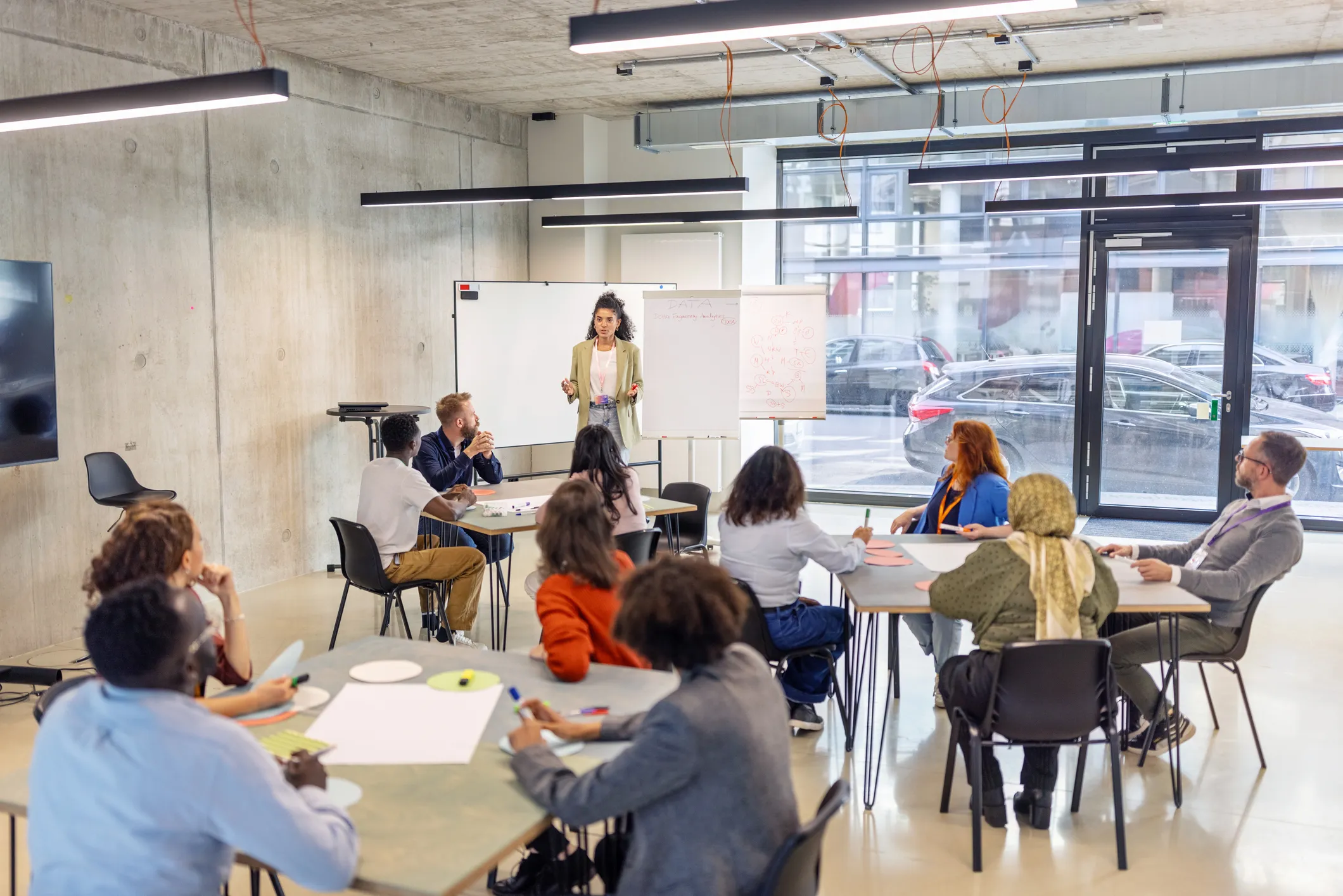 A group of adults sit at tables in a classroom-style setting while a presenter stands at the front near a whiteboard and flip chart.