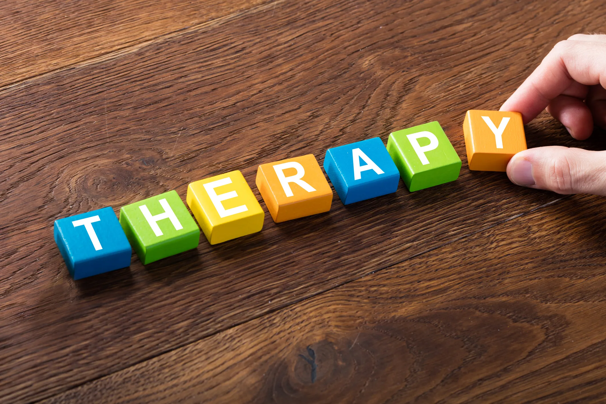 Colorful letter blocks spelling "THERAPY" on a wooden surface, with a hand placing the last block