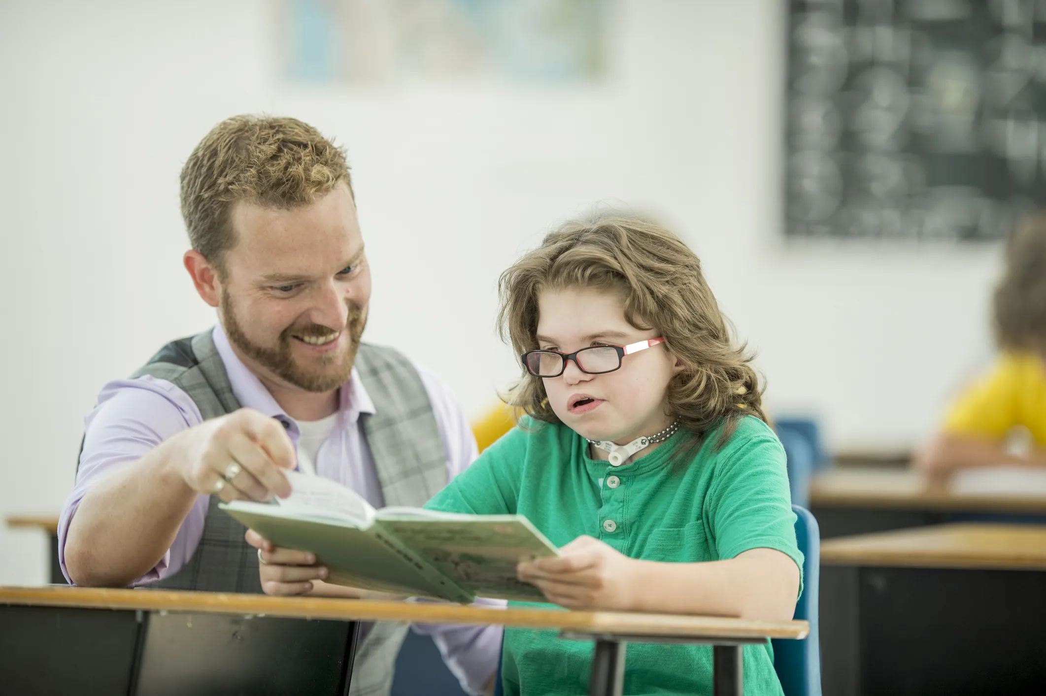 A Special Needs Assistant seated at a classroom desk reads a book with a child. The child is holding the book open while the Special Needs Assistant beside them points to the page. A chalkboard and other students are visible in the blurred background.